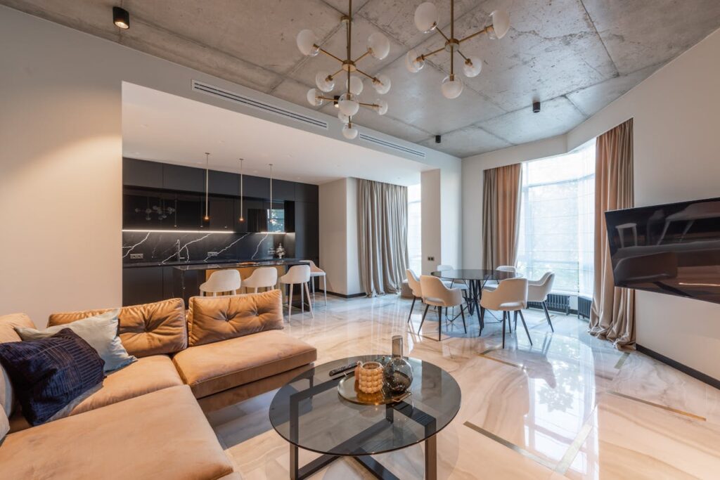 Interior of contemporary flat with sofa with pillows near kitchen with counter and round table with chairs near TV under chandelier near windows with curtains